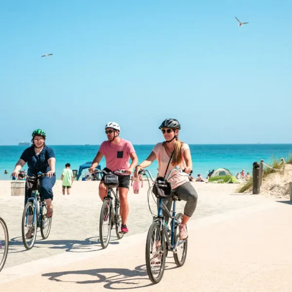 Vier personen fietsen met een helm op vlak voor een strand met heldere blauwe lucht in de achtergrond.